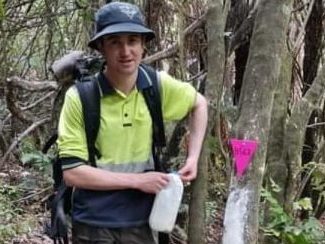 A young person in work wear stands by a tree with a bait station attached and a lure of flavoured flour to tempt the possums