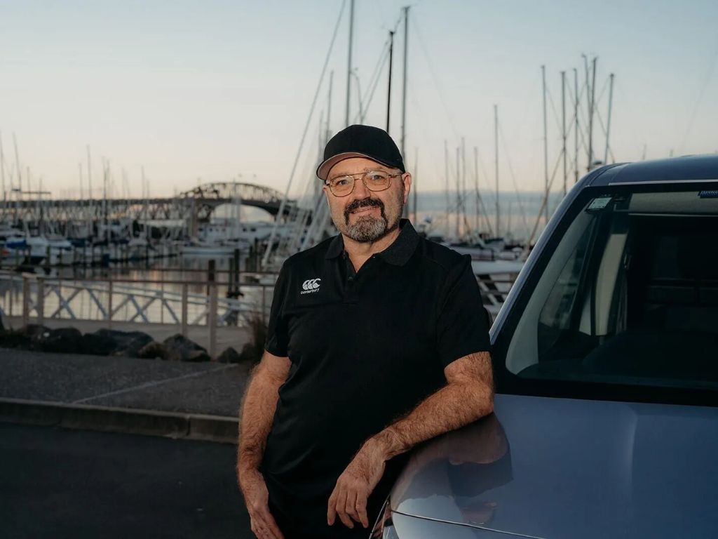 Owen leaning on the bonnet of his car, in front of the Auckland Harbour Bridge.