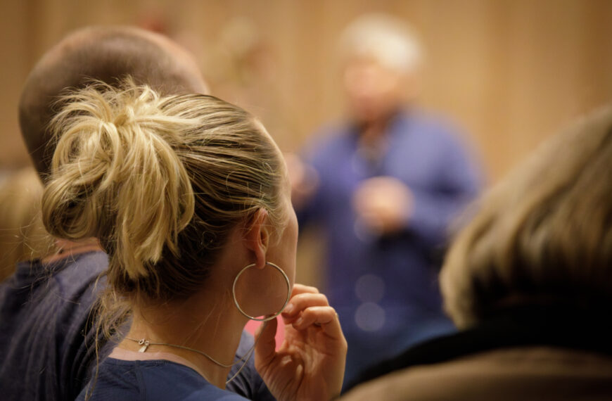 A woman with blonde hair is listening to a person in the background.