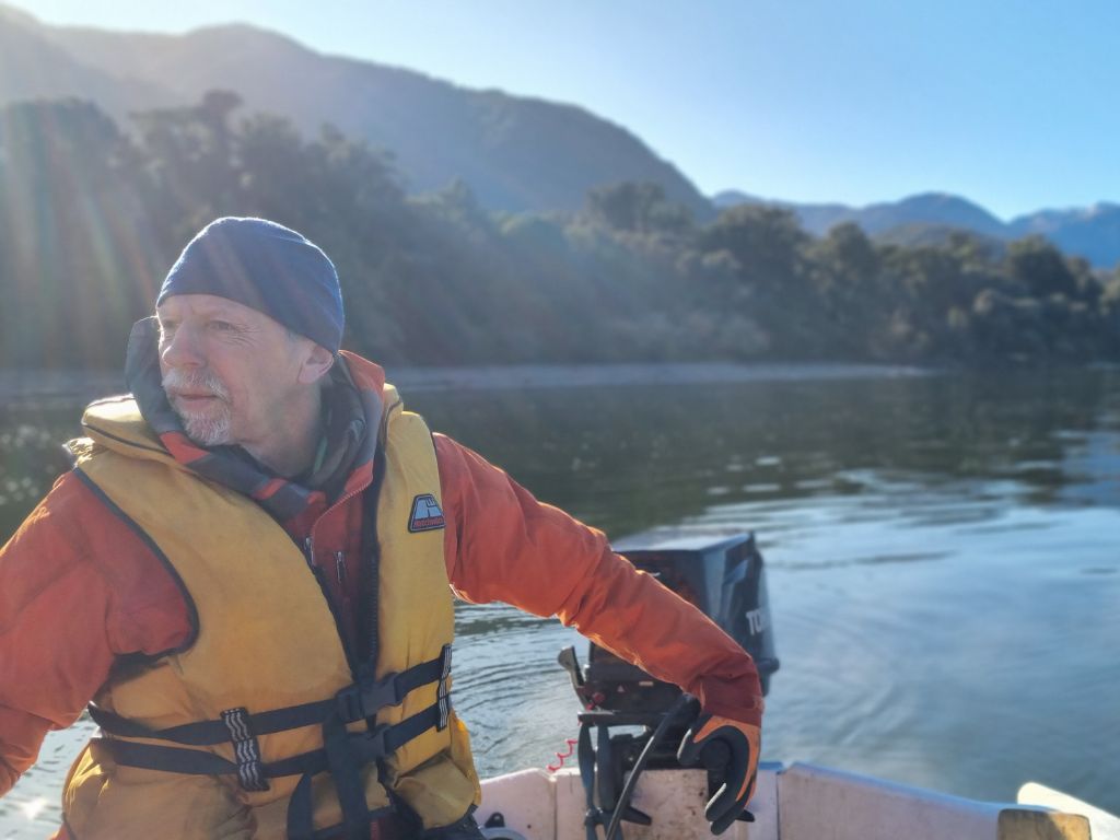 Lindsay driving a dinghy on the water, Fiordland bush in the background.