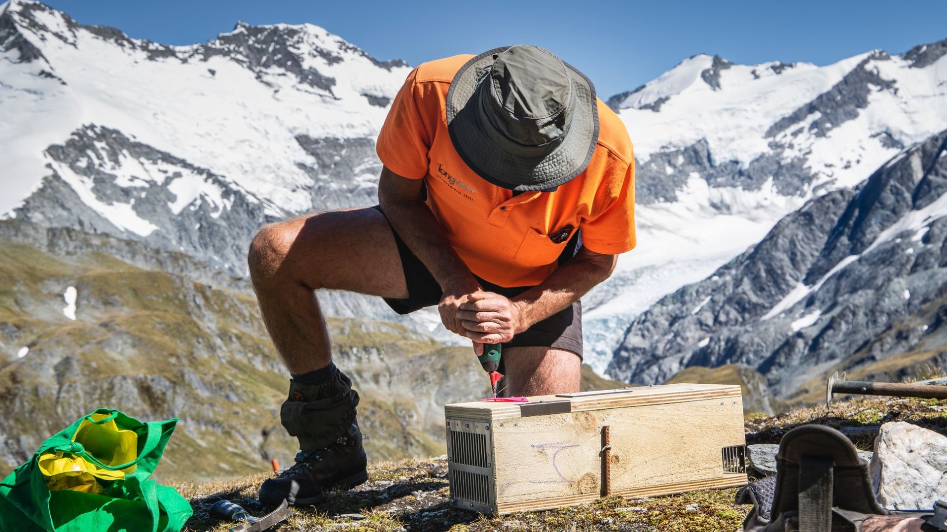 A man opening a trap box on a ridge with mountains in the background.