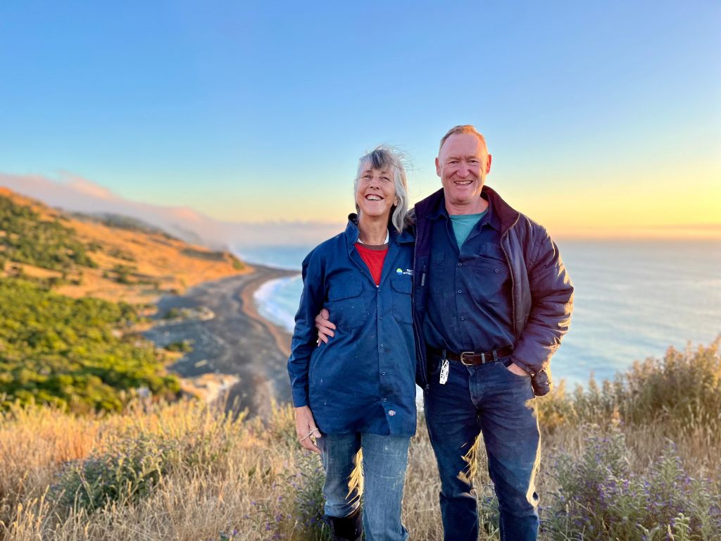 A couple standing on farmland, sea in the background.