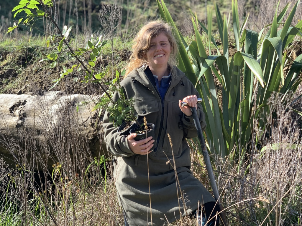 Miranda wells standing in an area for restoration. She is holding plants in one hand and a shovel in the other.
